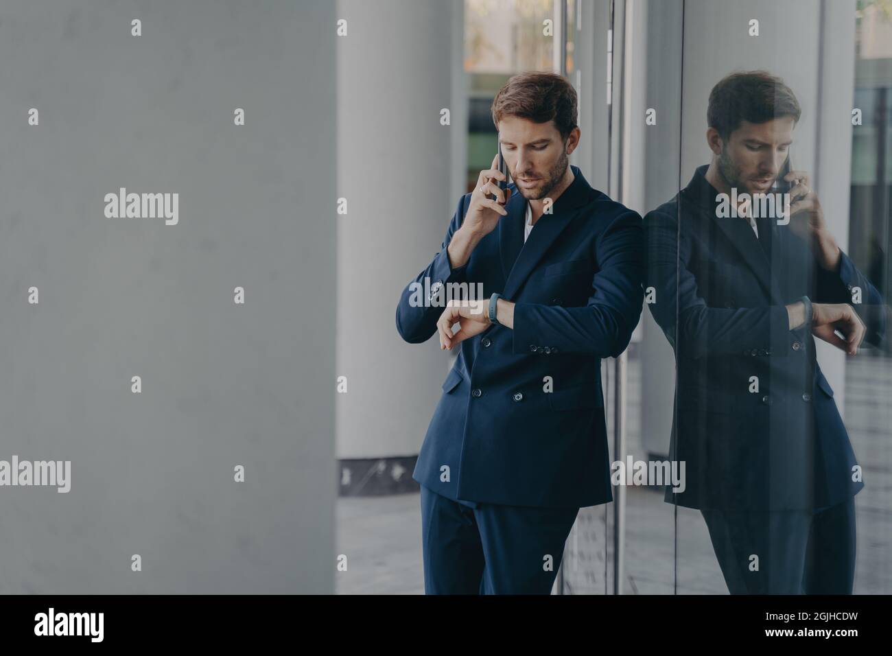 Nachdenklicher Geschäftsmann im Anzug, der mit dem Mobiltelefon telefoniert und draußen auf seine Handuhr schaut Stockfoto