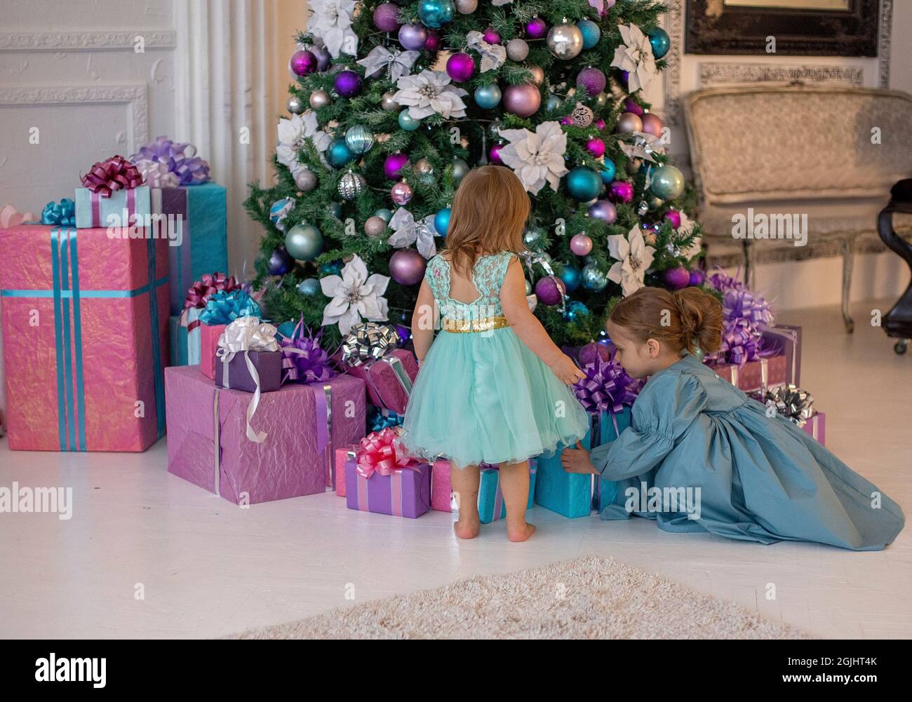 Zwei Mädchen in ausgefallenen Kleidern suchen Weihnachtsgeschenke unter einem wunderschön dekorierten Baum. Stockfoto