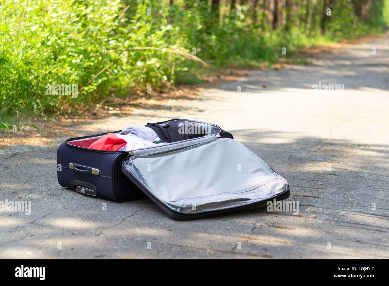 Ein offener Koffer mit Dingen liegt an einem sonnigen Sommertag auf einer Waldstraße vor einem Hintergrund von grünem Laub. Selektiver Fokus Stockfoto