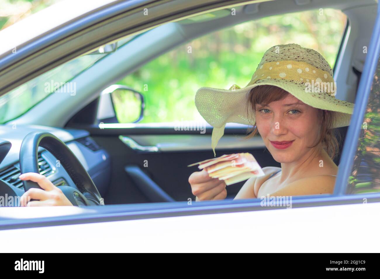 Ein Mädchen in einem Strohhut hält Geld aus, während es an einem sonnigen Sommertag in einem weißen Auto hinter dem Steuer sitzt. Selektiver Fokus Stockfoto