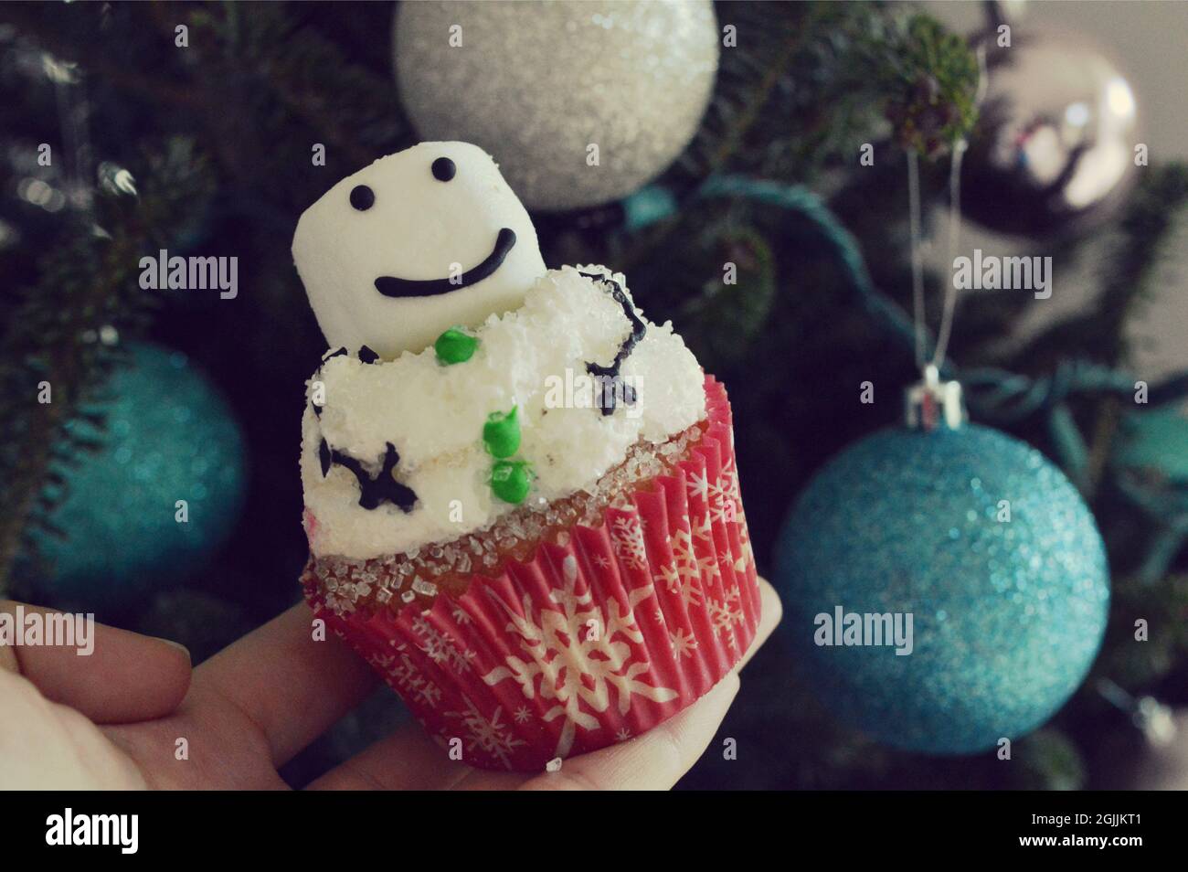 Hausgemachter Cupcake in Form eines Schneemanns vor dem Hintergrund eines Weihnachtsbaums, der mit Ornamenten verziert ist Stockfoto