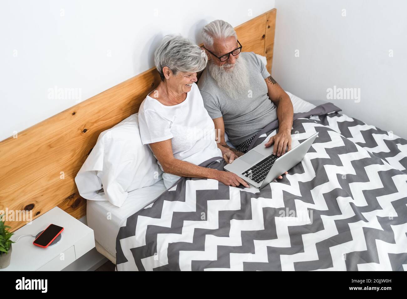 Glückliches Seniorenpaar, das Spaß beim Computergucken hat, während es unter den Decken im Bett lag - Technologie und Lifestyle-Konzept für ältere Menschen Stockfoto