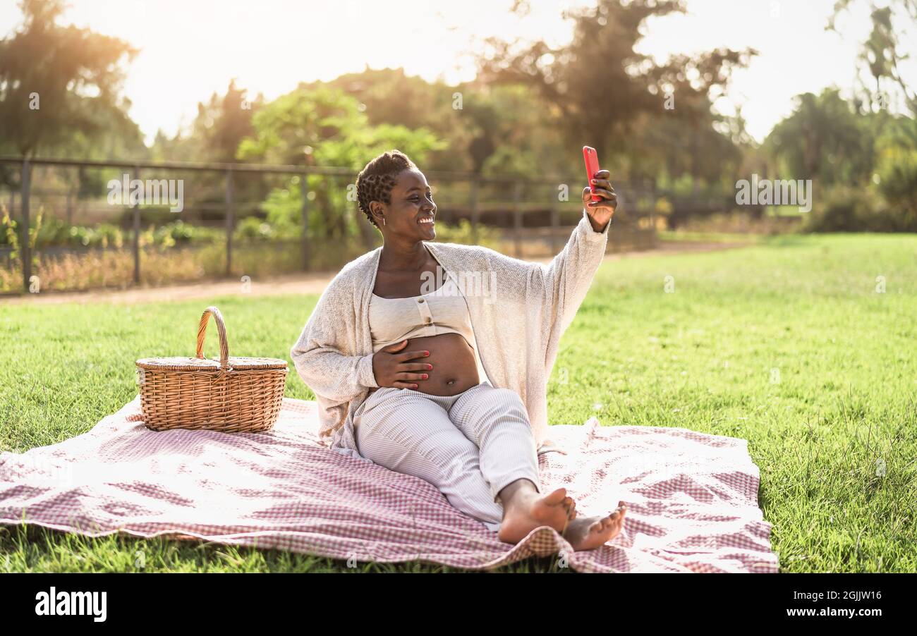 Afrikanische Schwangere, die Selfie mit dem Handy macht, während sie ein Picknick im Park macht - Mutterschaft Lifestyle-Konzept Stockfoto