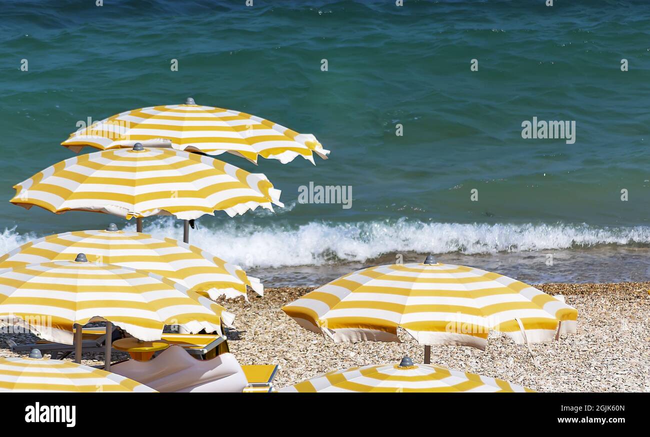 Gelb-weiß gestreifte Sonnenschirme öffnen sich an einem Kieselstrand an einem windigen Sommertag. Meereswellen im Hintergrund. Urlaub und Entspannung Stockfoto
