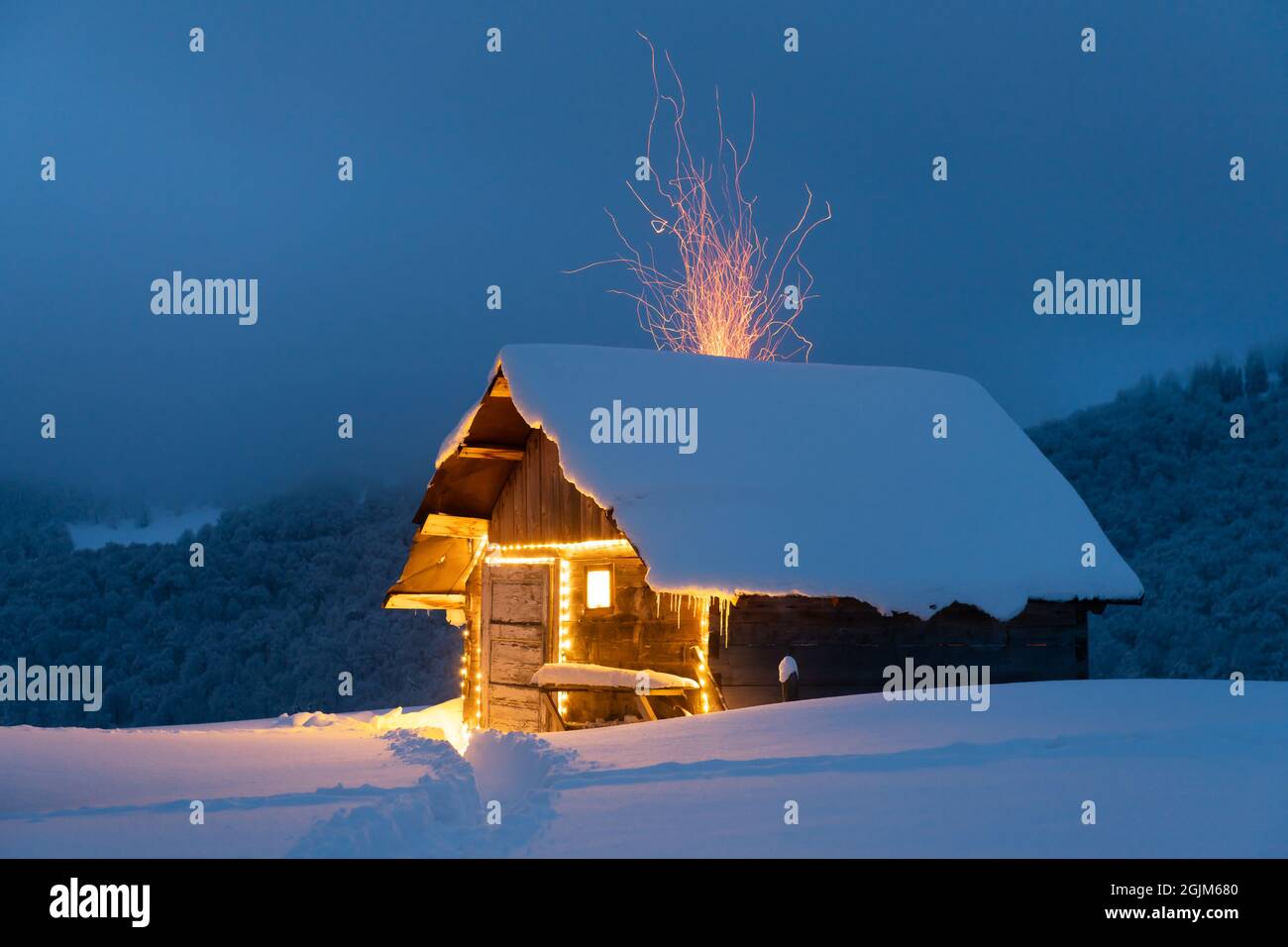 Fantastische Winterlandschaft mit glühender Holzhütte im verschneiten Wald. Feuerfunken fliegen aus dem Kamin. Weihnachtsfeiertagskonzept Stockfoto