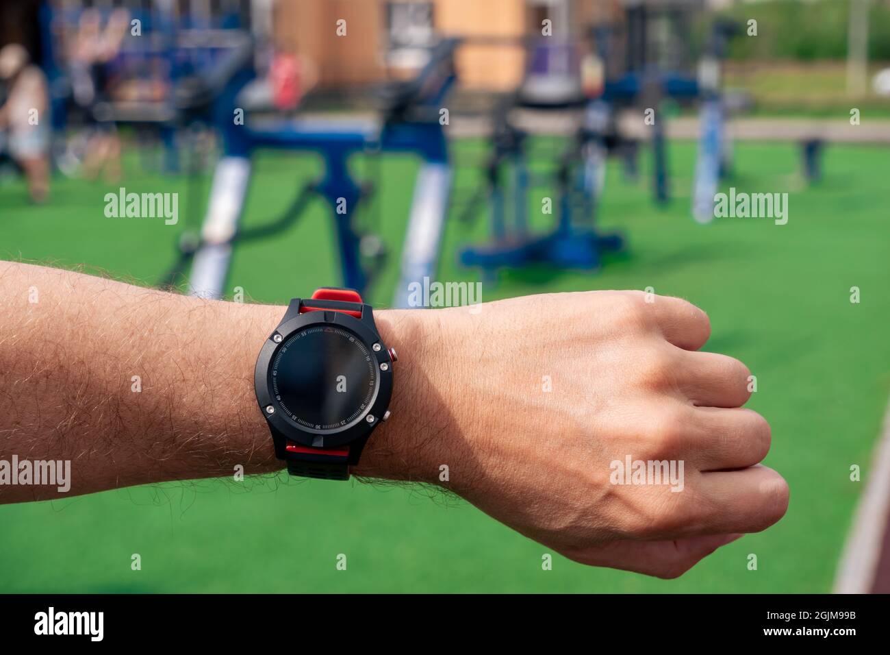 Schauen Sie sich eine Smartwatch an, die auf der Hand eines Mannes vor dem Hintergrund von Fitnessgeräten im Freien steht Stockfoto