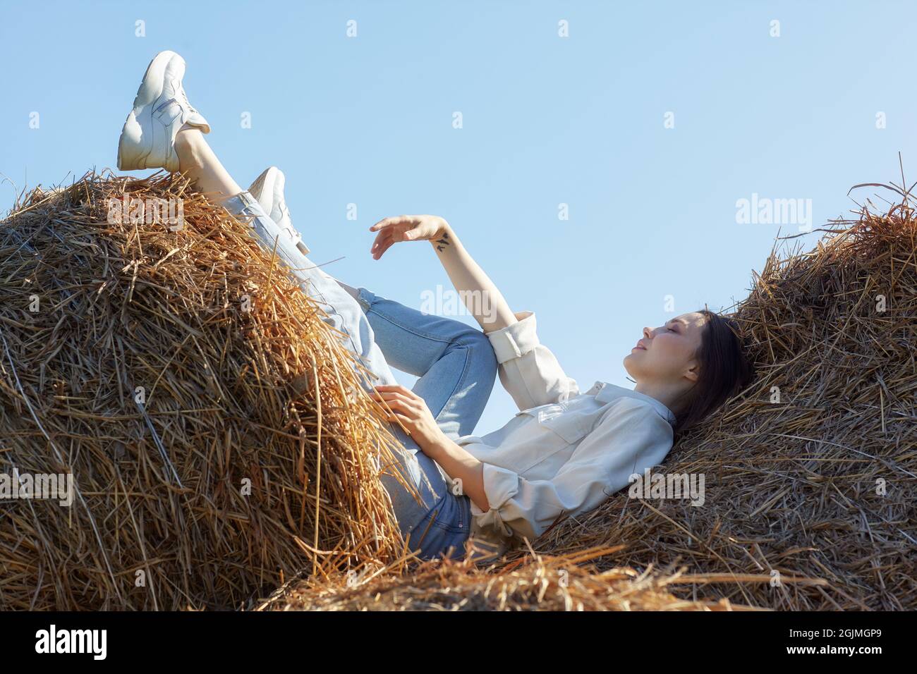 Junge Brünette Frau in einem weißen Hemd und Jeans liegt in den Rollen von Heu. Romantisches Landmädchen auf Heu. Stockfoto