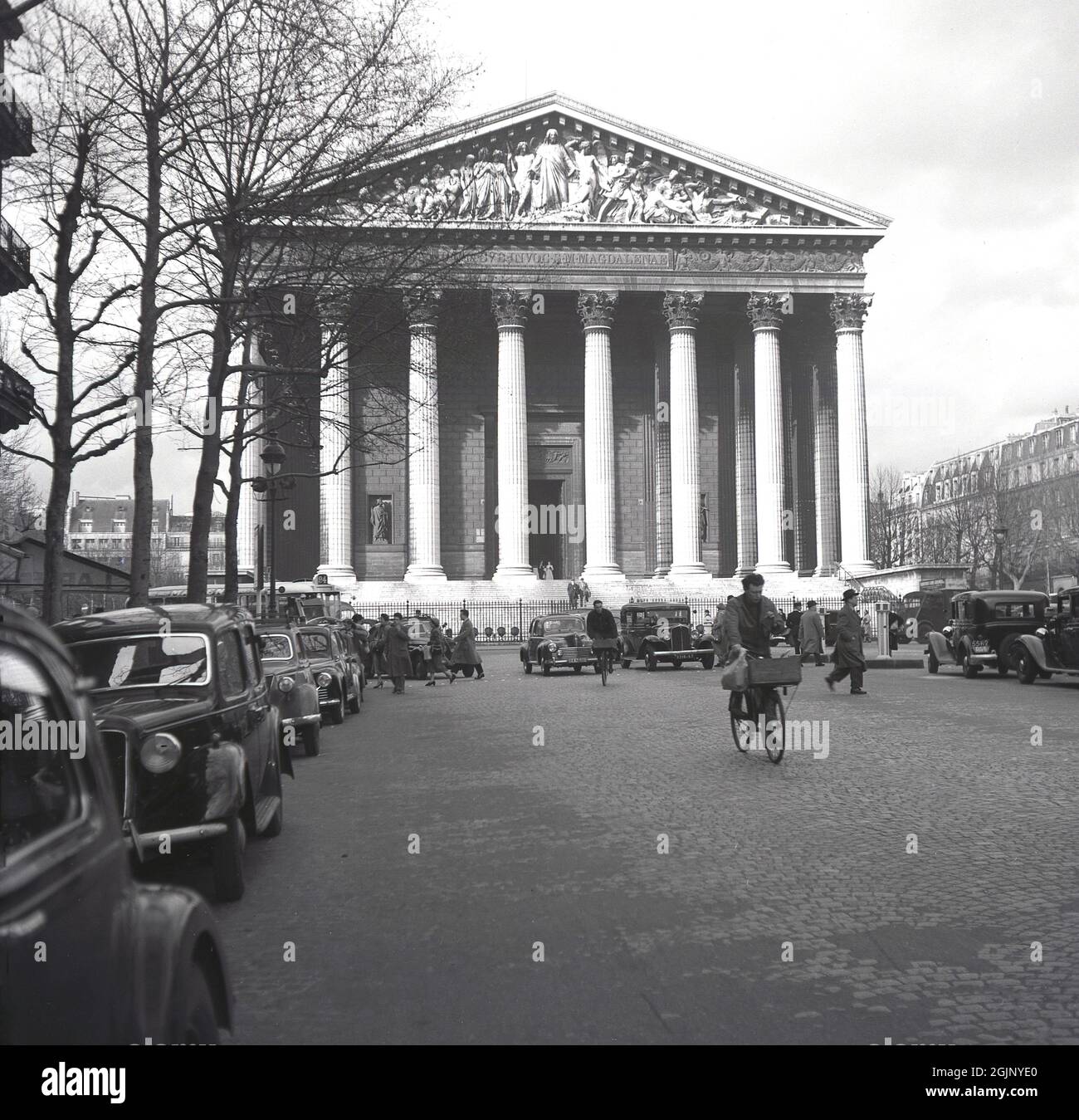 1950er Jahre, historische Ansicht der katholischen Kirche, La Madeleine mit Autos aus der Zeit auf der gepflasterten Straße geparkt und zwei Männer auf Lieferfahrrädern mit vorderen Körben, Paris, Frankreich. Erbaut als römischer Tempel mit seinen großen Säulen als Zeugnis des militärischen Ruhmes Napoleons, wurde er später nach Jesu Gefährtin, Maria Magdalena, benannt. Stockfoto