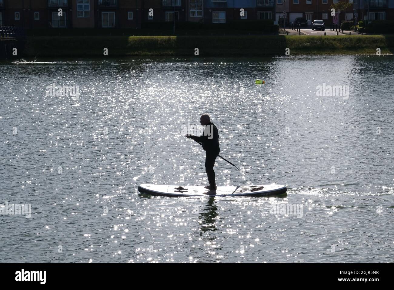 Ein männlicher Stand-up-Paddle-Boarder, der vor dem Licht mit spiegelbildlichen Reflexionen des Sonnenlichts auf der Wasseroberfläche am North Dock von Llanelli, heute eine Freizeitanlage, abgebildet ist Stockfoto