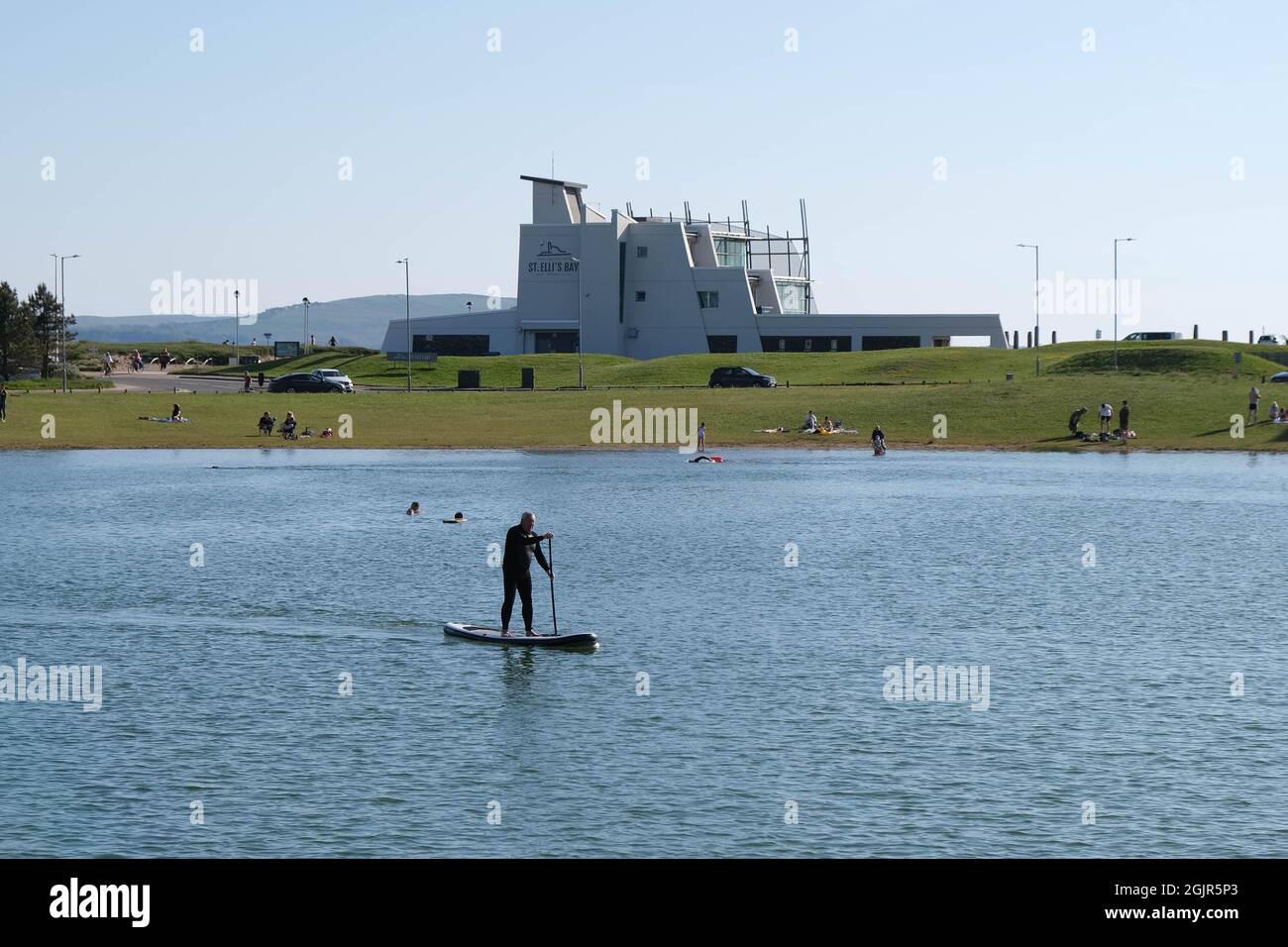 Ein männlicher Stand-up-Paddle-Boarder mitten im North Dock von Llanelli (jetzt ein Freizeitbad) mit St. Elli's Bay Cafe & Bistro im Hintergrund (ehemals Millennium Coastal Park Discovery Center), der auf der Südseite den Strand von Llanelli überblickt. Stockfoto