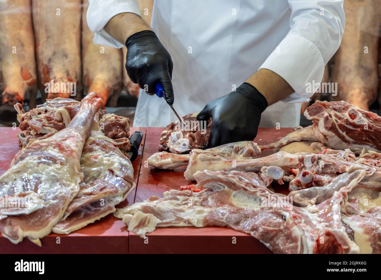 Metzgermann in schwarzen Handschuhen schneidet Lammfleisch in der Metzgerei. Koch Koch mit weißer Schürze schneidet Fleisch Stockfoto