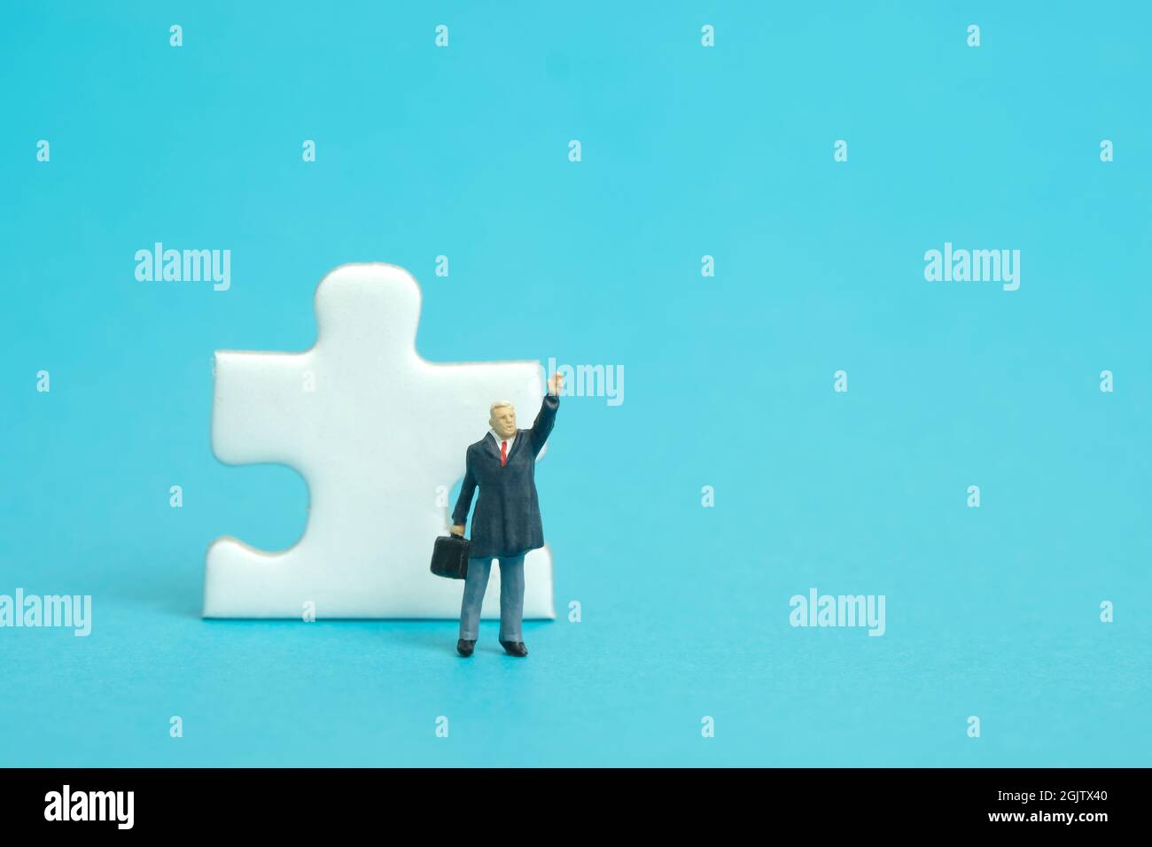 Miniatur Menschen Spielzeug Figur Fotografie. Ein Geschäftsmann, der vor einem Puzzle-Puzzleteil steht, während er seine Hand hebt. Isoliert auf blauem Hintergrund. Ima Stockfoto