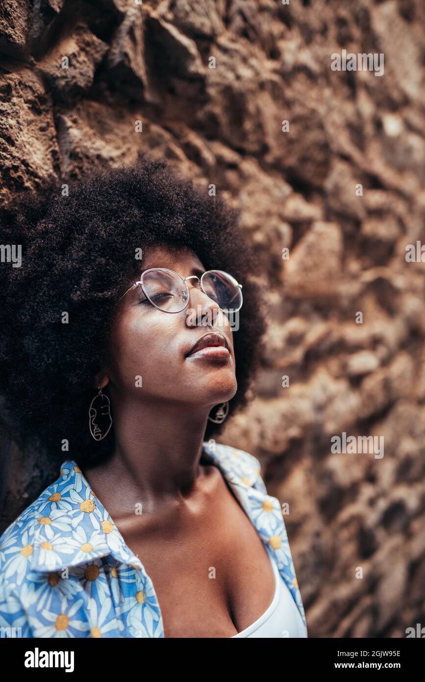 afro-Frau mit Brille und lässiger Kleidung mit geschlossenen Augen. Sie lehnt sich an eine Steinmauer. Konzept der Entspannung und Meditation Stockfoto