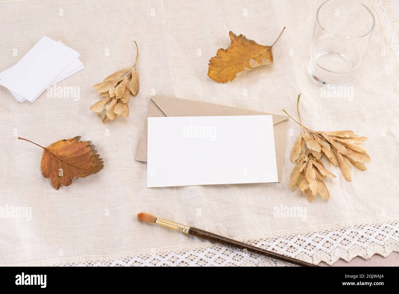 Eine Visitenkarte auf einem Umschlag, einem Brief oder einer Einladung vor dem Hintergrund einer hellen Leinenserviette mit abgefallenen Blättern. Hochzeit Schreibwaren Mockup Stockfoto