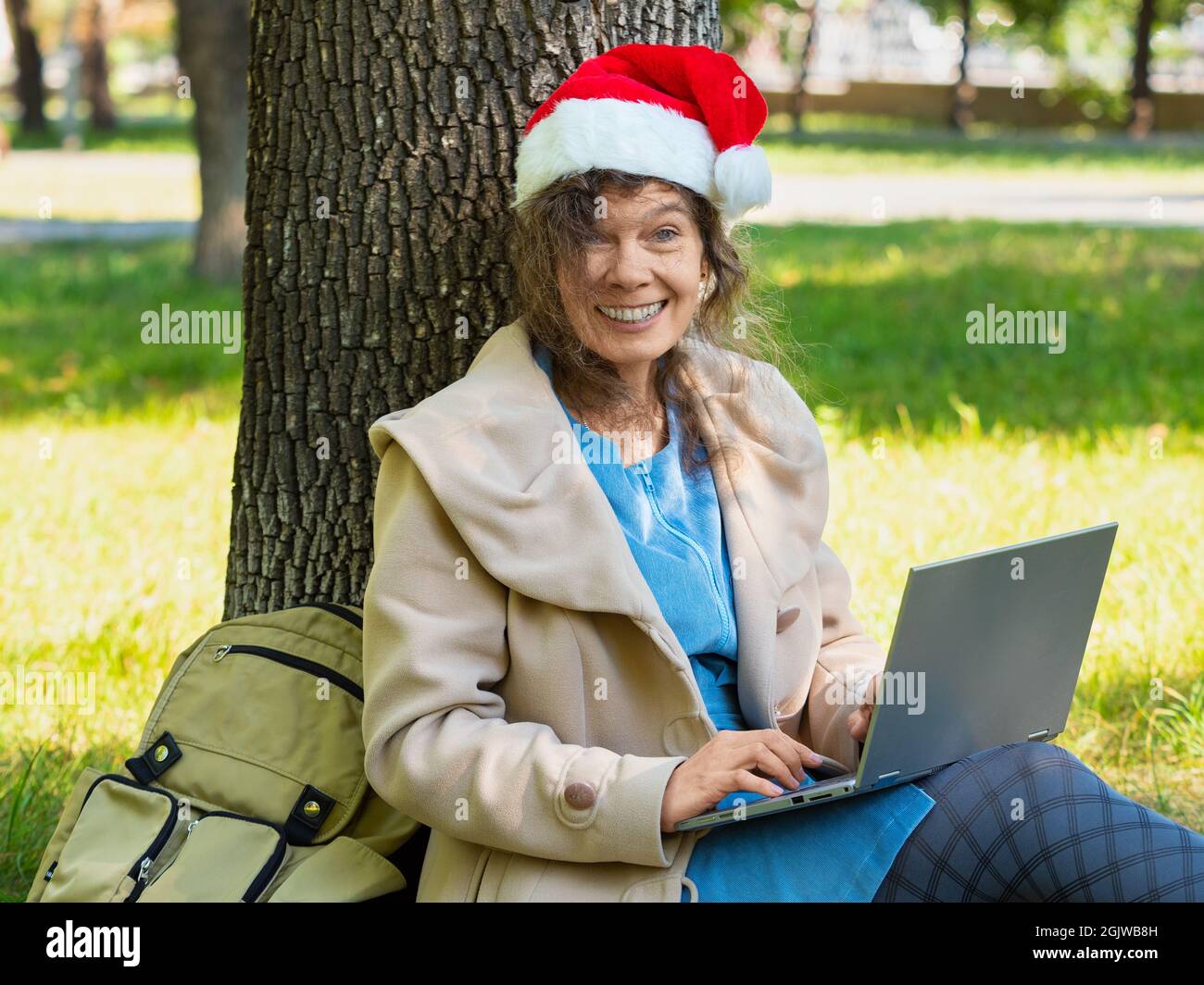 Foto einer Frau mittleren Alters in Santa hat mit Online-Videoanruf. Sie sitzt auf dem Gras unter dem Baum im Stadtpark mit Laptop. Sie lächelte und Stockfoto