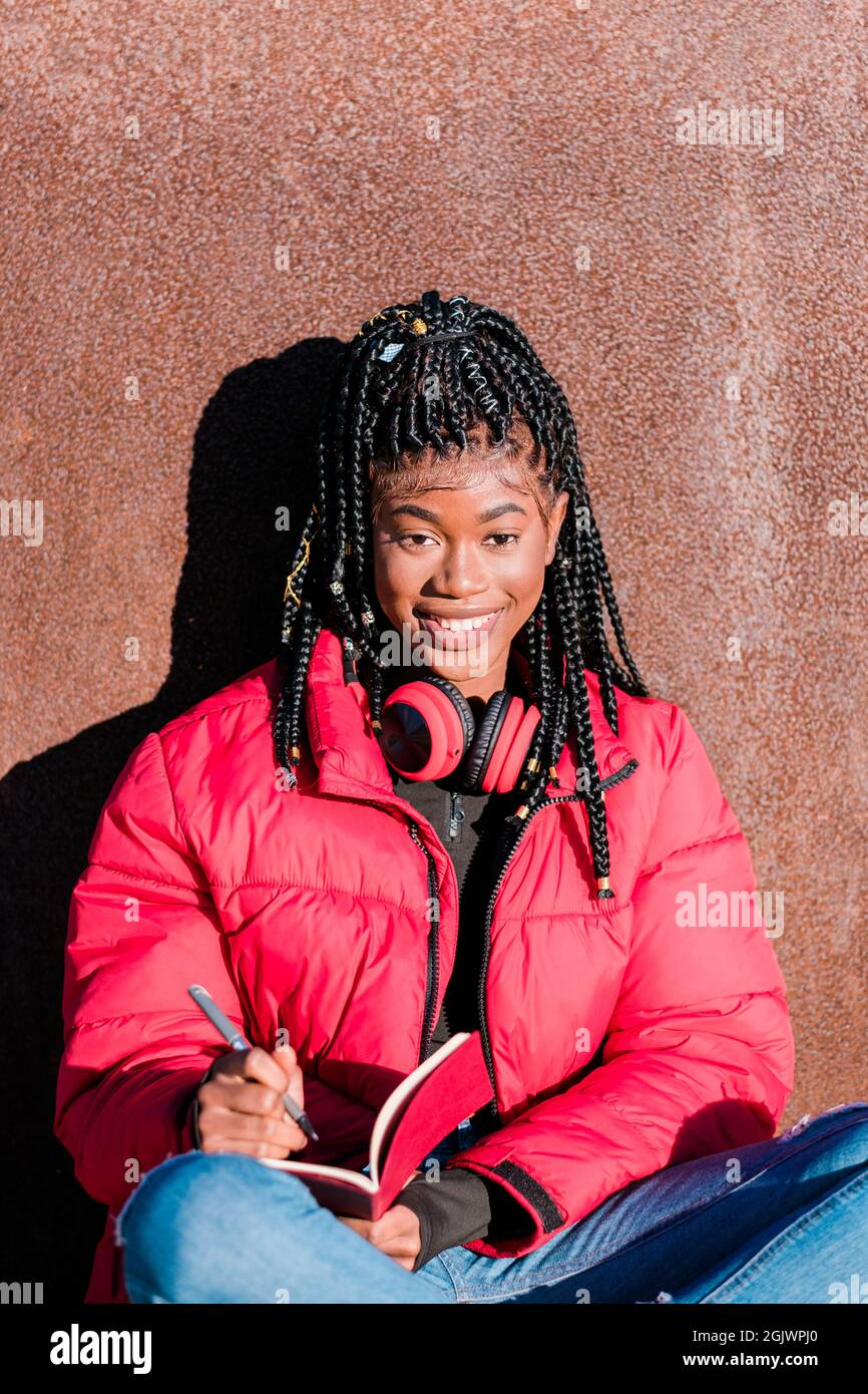 Schöne afrikanische Frau trägt rote Jacke mit Kopfhörern im Freien. Eine schwarze Frau, die in einem Notizbuch schreibt und die Kamera anschaut Stockfoto