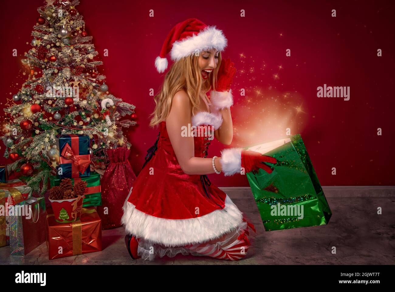 Nettes Mädchen als Weihnachtsmann gekleidet, Blick in einer Geschenktüte mit einem Glanz und Sterne, hinter dem Weihnachtsbaum und Geschenke. Stockfoto