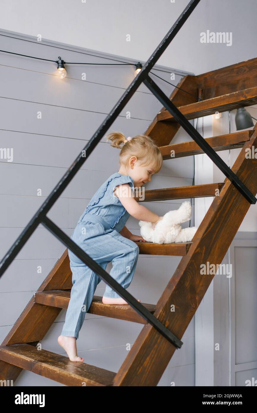 Ein süßes, zweijähriges Mädchen in einem Jeansjumpsuit mit einem weichen Spielzeug in den Händen steigt die Treppe hinauf in den zweiten Stock des Hauses Stockfoto