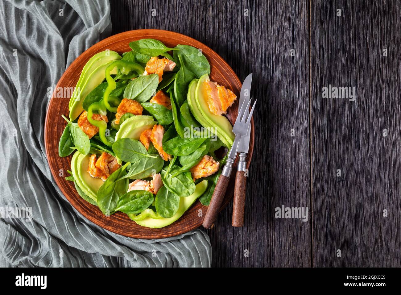 Lachs Spinat Avocado und grüner Pfeffer Salat auf einem braunen rustikalen Teller mit Gabel und Messer auf einem Mooreichentisch, mediterrane gesunde Ernährung, flaches Lay, Fr. Stockfoto
