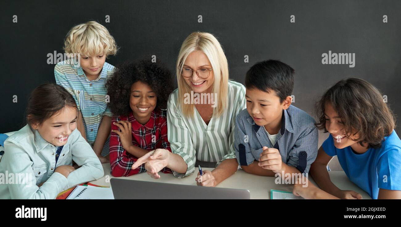 Fröhlicher Lehrer, der mit verschiedenen Schulkindern am Schreibtisch mit einem Laptop zusammensitzt. Stockfoto