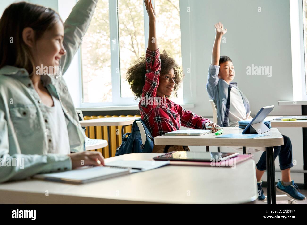 Verschiedene Schulkinder, die am Schreibtisch sitzen und beim Unterricht die Hände heben. Stockfoto