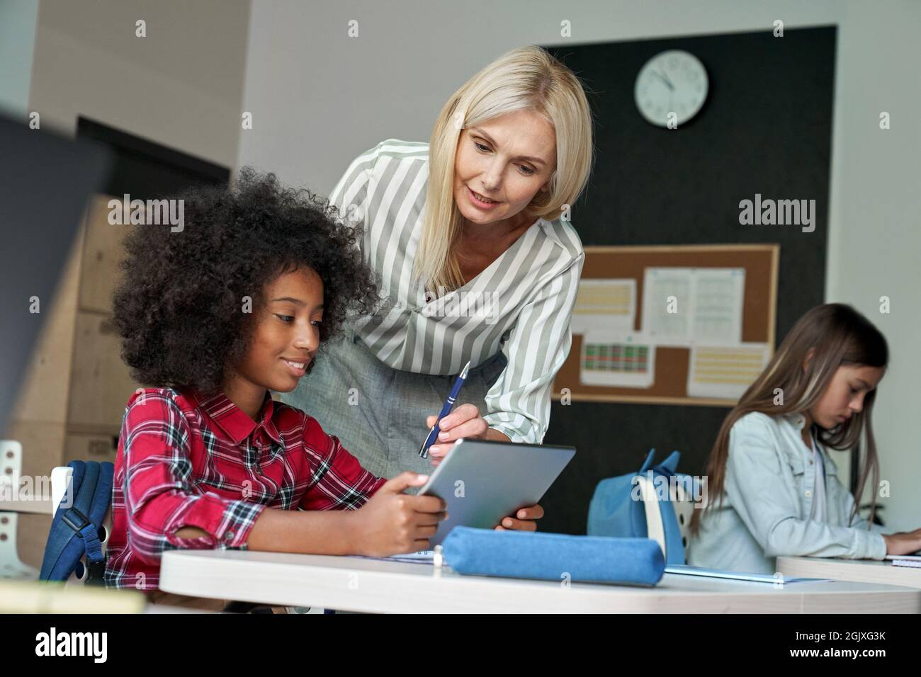 Fröhliches afroamerikanisches Schulkind hört dem Lehrer zu, der im Klassenzimmer ein Tablet hält. Stockfoto