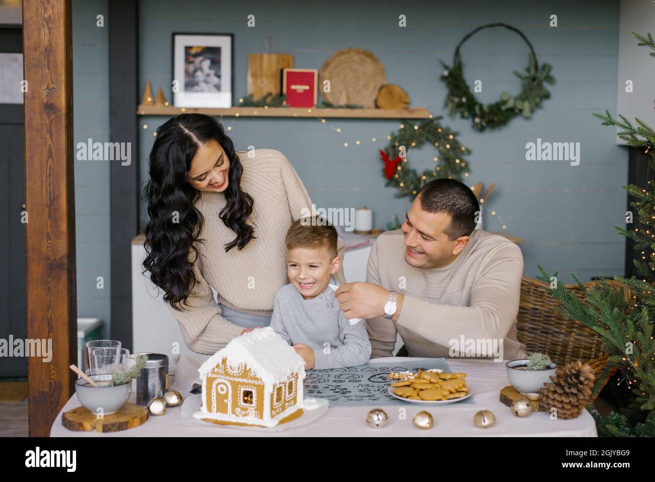 Die Familie backt zu Weihnachten ein Lebkuchenhaus mit Süßigkeiten, der Vater legt seinem Sohn das Sahnehäubchen auf die Nase, sie lachen Stockfoto