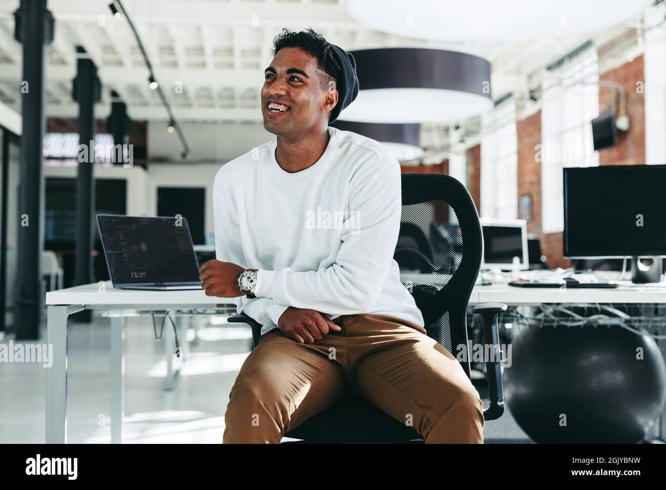 Glücklicher Software-Entwickler lächelt in einem modernen Büro. Selbstbewusster junger Geschäftsmann, der alleine dasitzt und wegschaut. Junger Geschäftsmann, der eine Pause einnahm Stockfoto