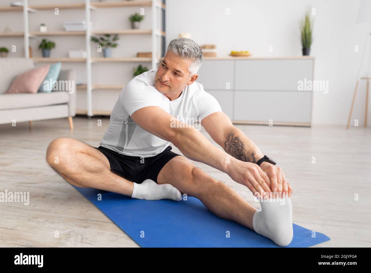 Aktiver Lebensstil. Sportlicher reifer Mann, der sich das Bein streckt, auf einer Yogamatte im Wohnzimmer arbeitet, Platz für Kopien Stockfoto