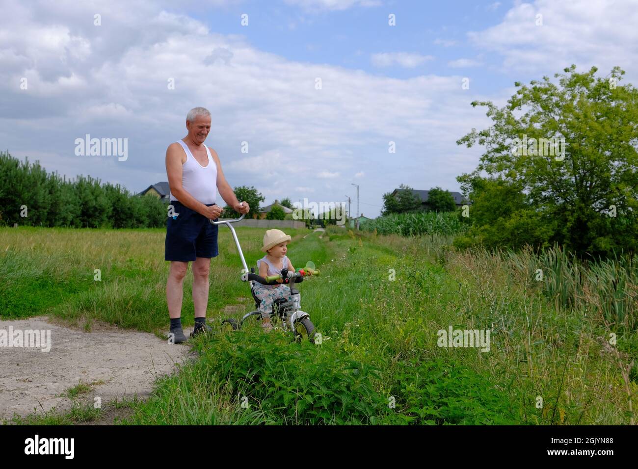 In voller Länge Ansicht eines älteren Mannes mit einem niedlichen kleinen Mädchen, das auf der grünen Wiese spazierengeht. Das kleine Mädchen sitzt auf dem Fahrrad auf dem grünen Gras Stockfoto