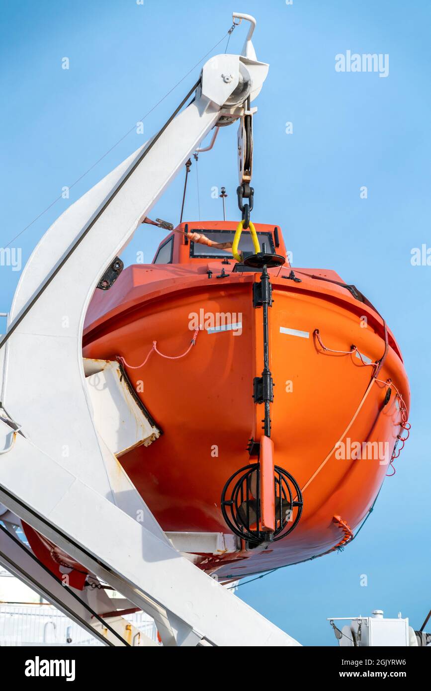 Vertikale Aufnahme eines orangefarbenen Rettungsbootes, das an einem Kran an Bord eines Segelschiffs hängt, mit blauem Himmel im Hintergrund. Stockfoto
