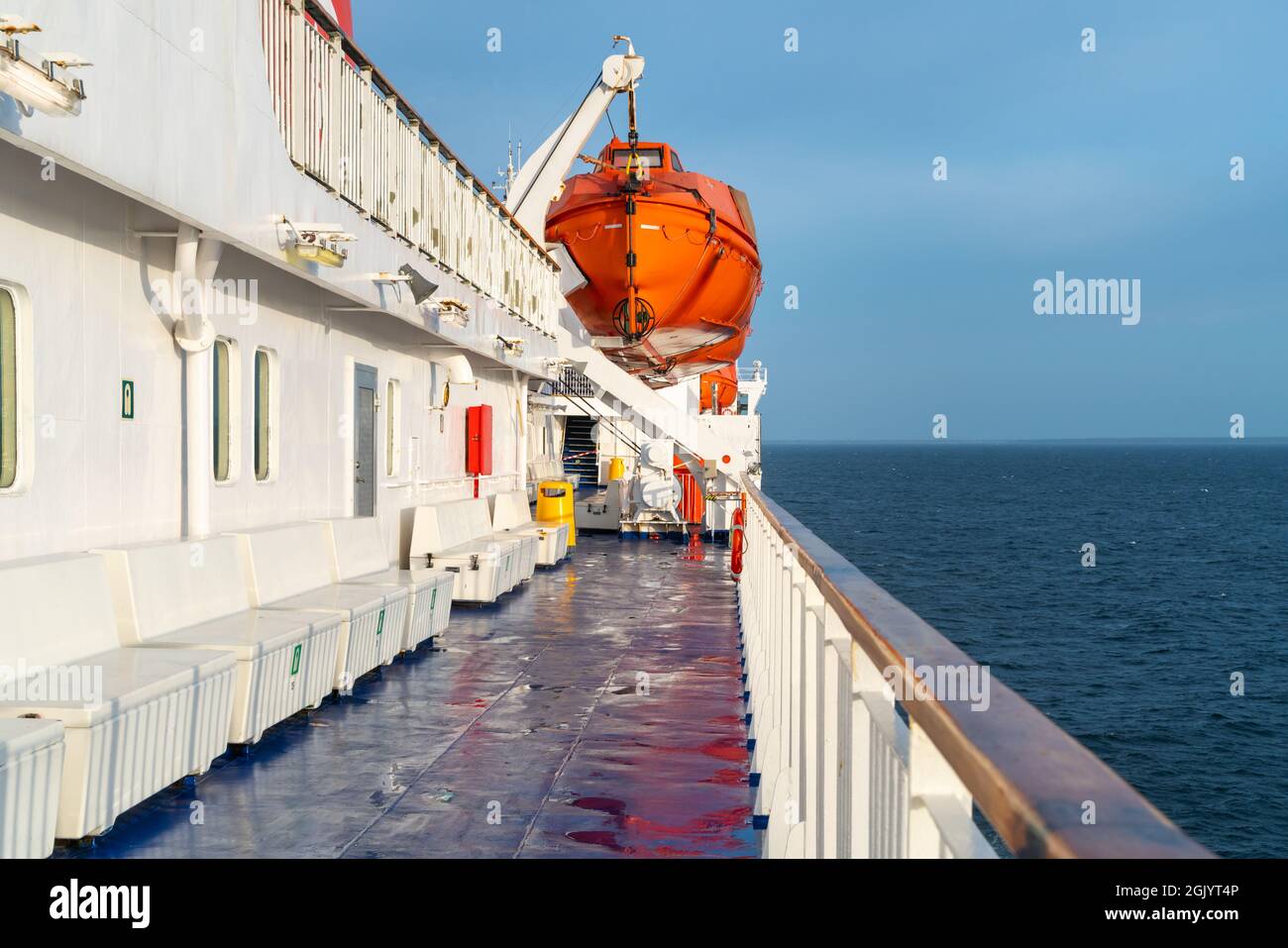 Orange Life Boot hängt an einem Kran auf einem Deck des Segelschiffs mit Ozeanhorizont im Hintergrund und Holzgeländer vor Stockfoto