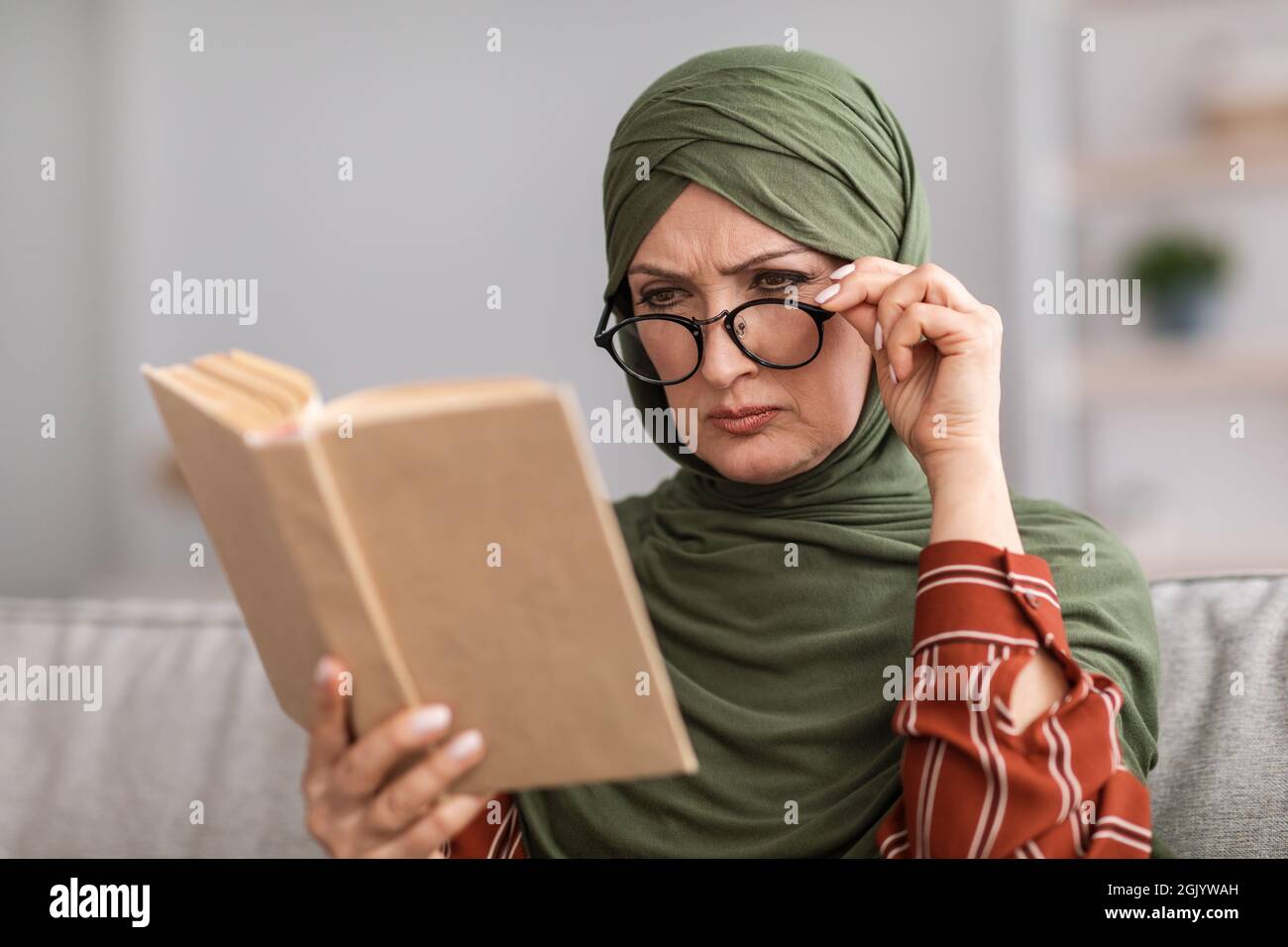 Ältere Muslimische Frau Mit Schlechtem Sehvermögen Beim Lesen Des Buches Zu Hause Stockfoto