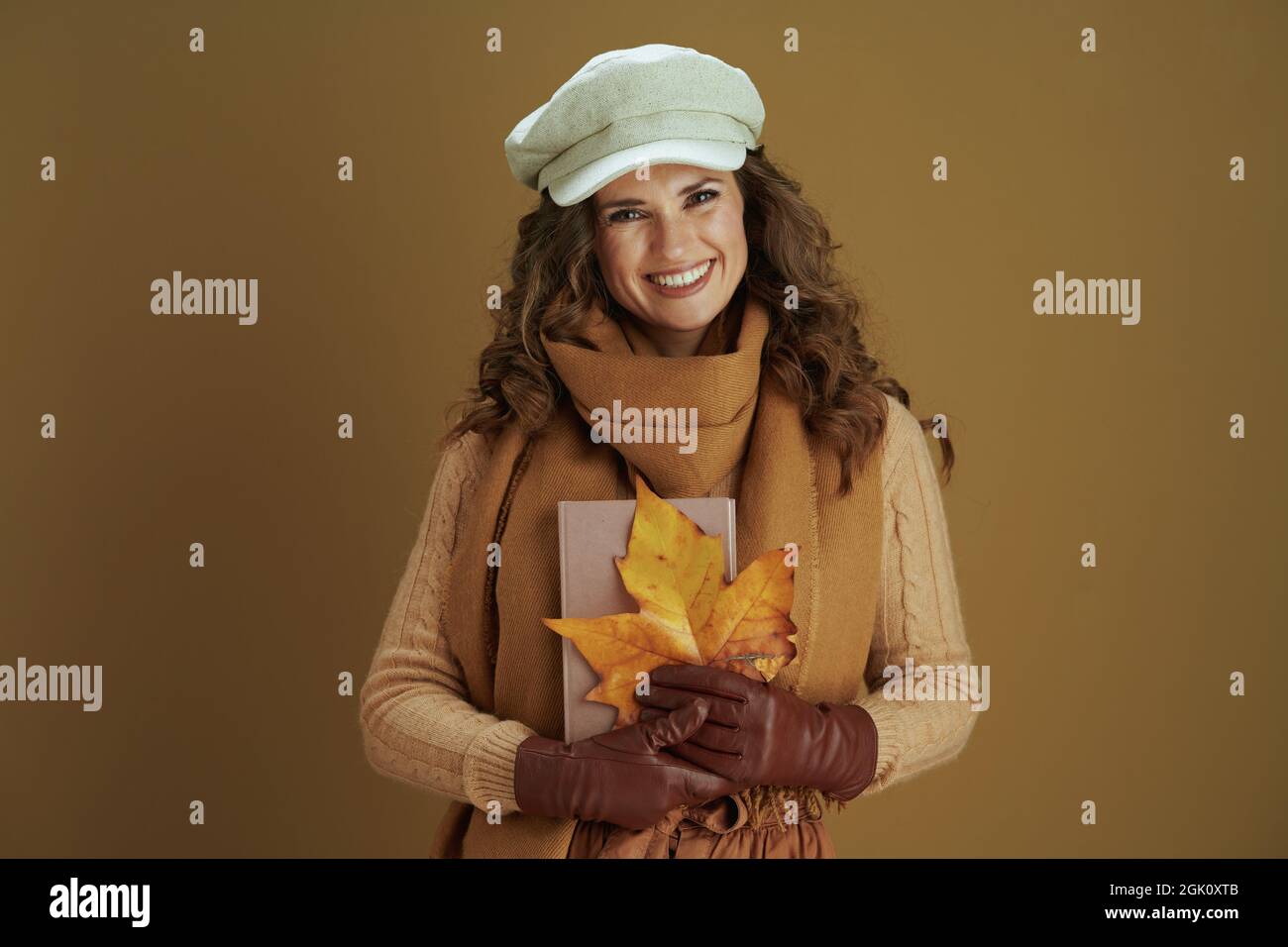 Hallo september. Portrait einer fröhlichen trendigen Hausfrau im Schal mit gelbem Ahornblatt, Buch und Lederhandschuhen auf braunem Hintergrund. Stockfoto