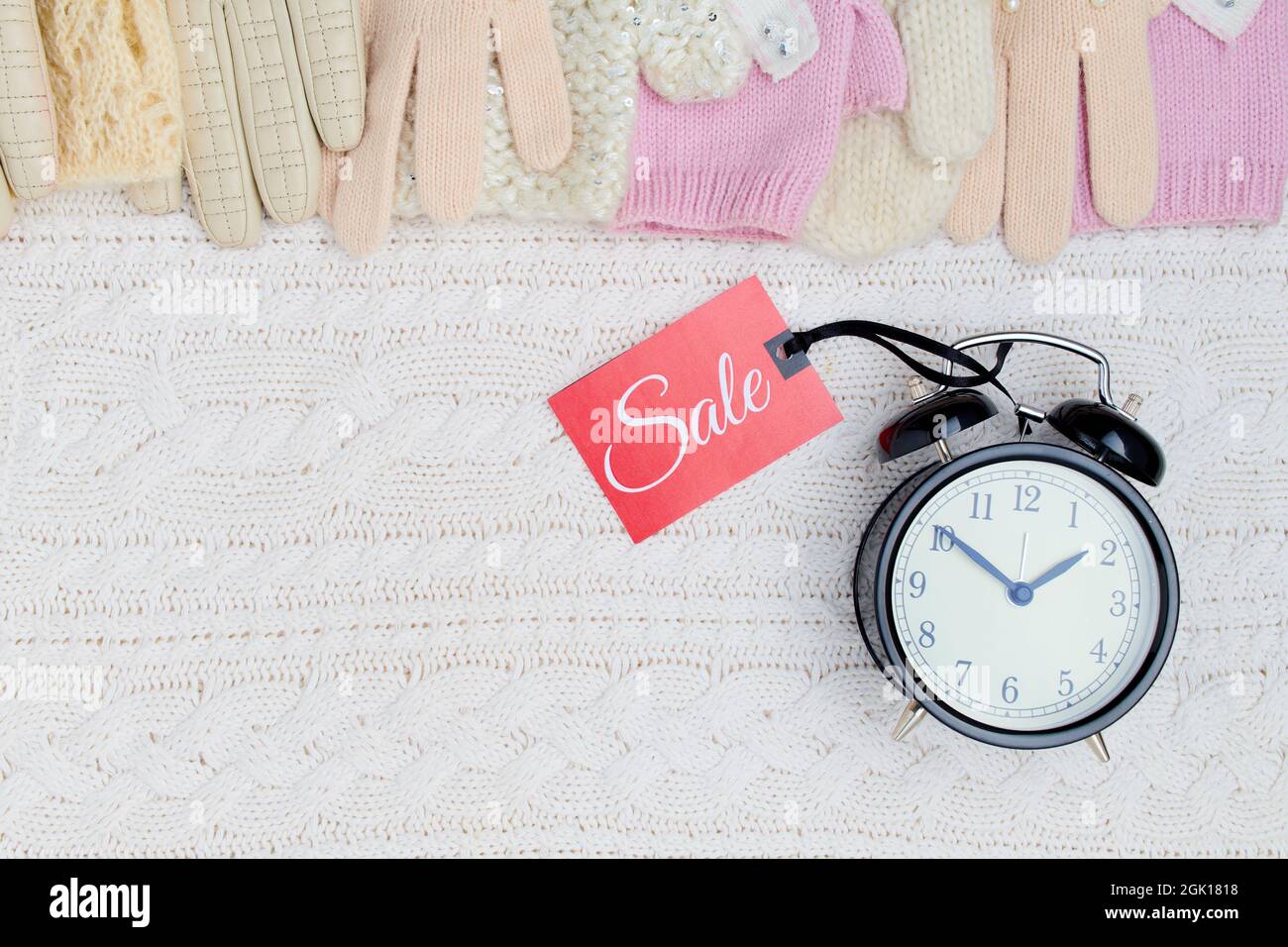 Winter-Flat-Lay mit Wecker, Handschuhen und Sale-Tag auf weißem gestricktem Hintergrund. Stockfoto