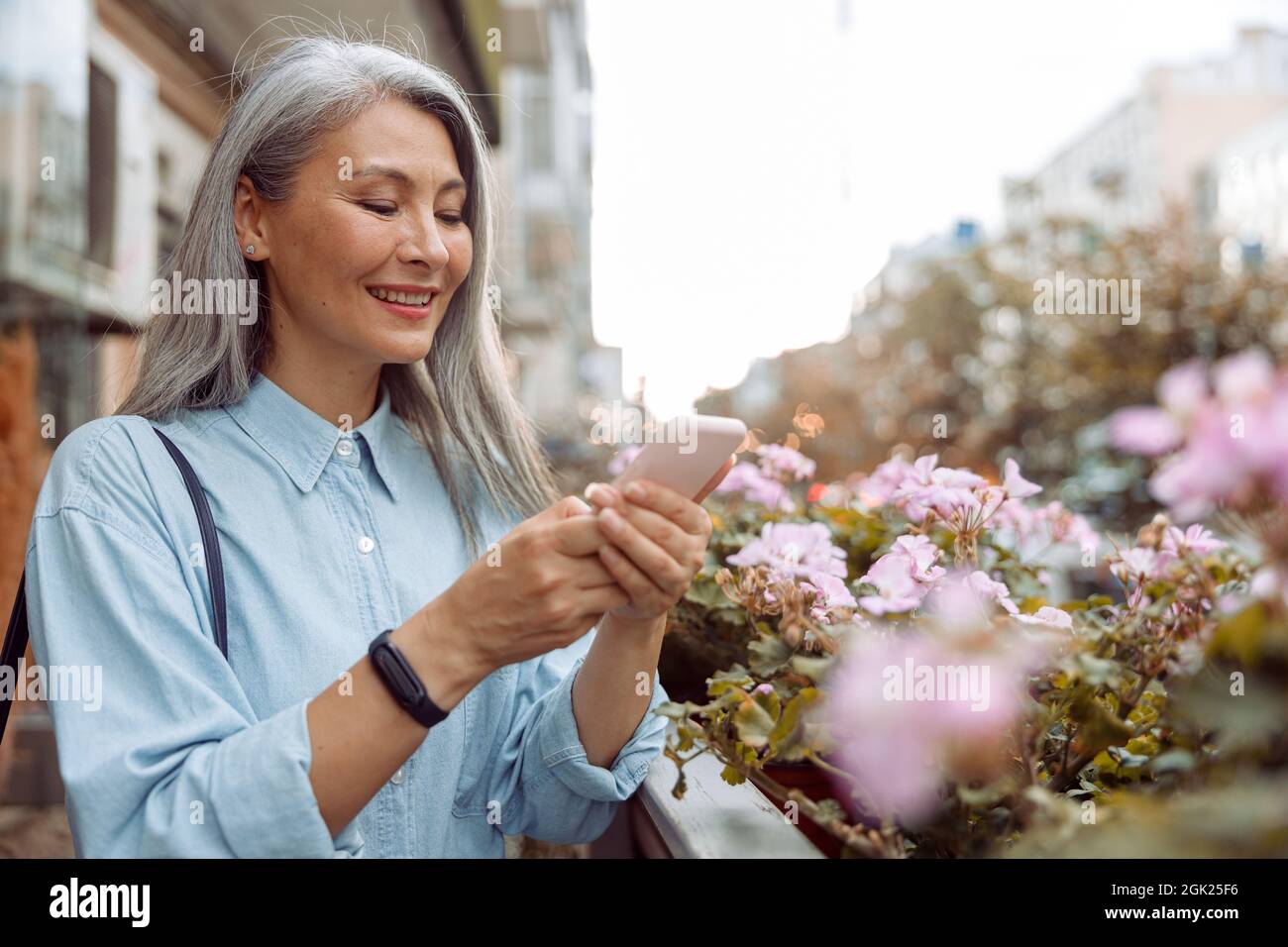 Lächelnde grauhaarige asiatische Frau benutzt Handy, das auf der Außenterrasse steht Stockfoto