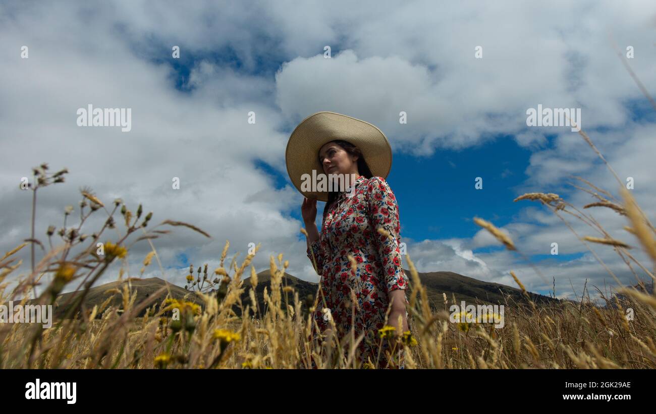 Glückliche junge Frau in Blumenkleid und langem Hut von unten betrachtet, die an einem bewölkten Tag mit blauem Himmel und Bergen mitten in einem Weizenfeld spazieren geht Stockfoto