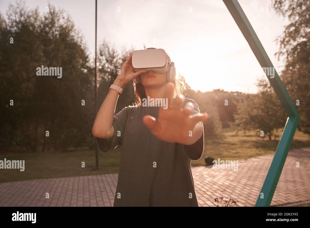 Nahaufnahme eines Mädchens, das mit den Händen eine Virtual-Reality-Brille auf dem Kopf hält und auf einem Hintergrund von grünen Bäumen steht. Er reicht eine Hand an die Kamera. Hochwertige Fotos Stockfoto