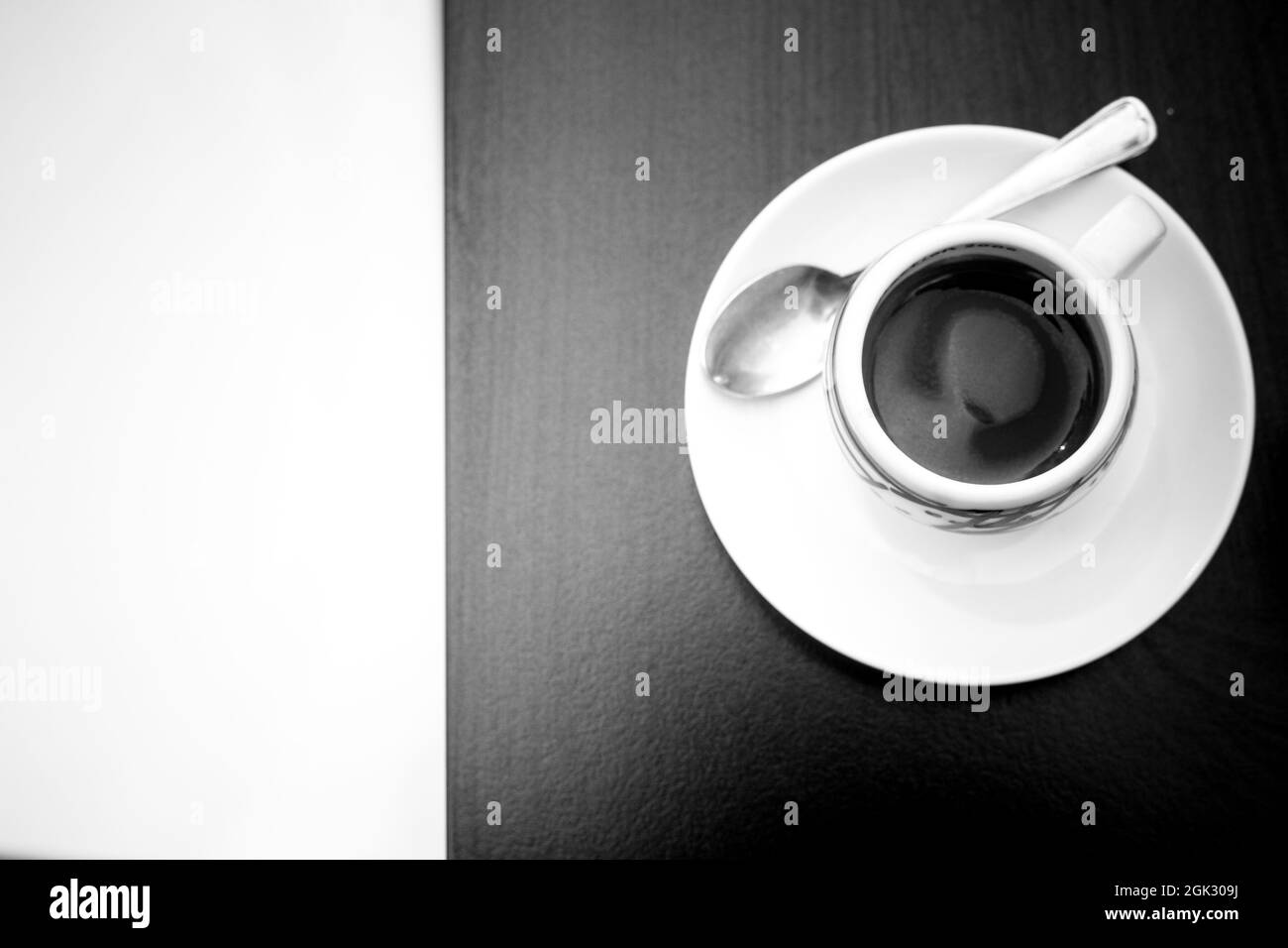 Monochromes Bild der Draufsicht auf eine Kaffeetasse mit Teller und Teelöffel auf einem dunklen Tisch und Kopierraum auf der linken Seite. Geometrisches Espresso-Konzept. Kaffee Stockfoto