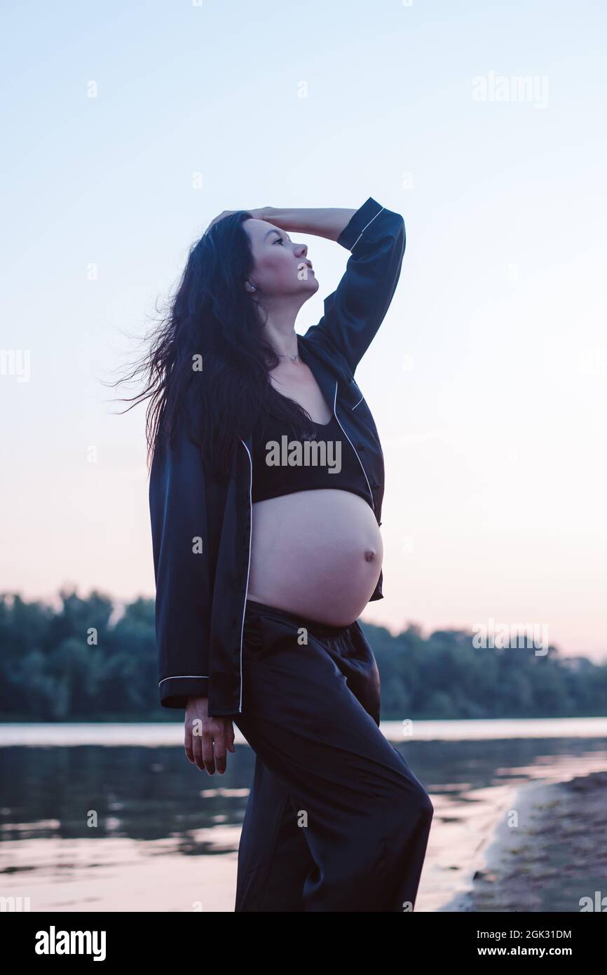Brunette posiert vor dem Hintergrund der Natur. Attraktive junge Schwangerin in dunklen Seide Pyjamas posiert und berührt ihre langen braunen Haare weht in Stockfoto