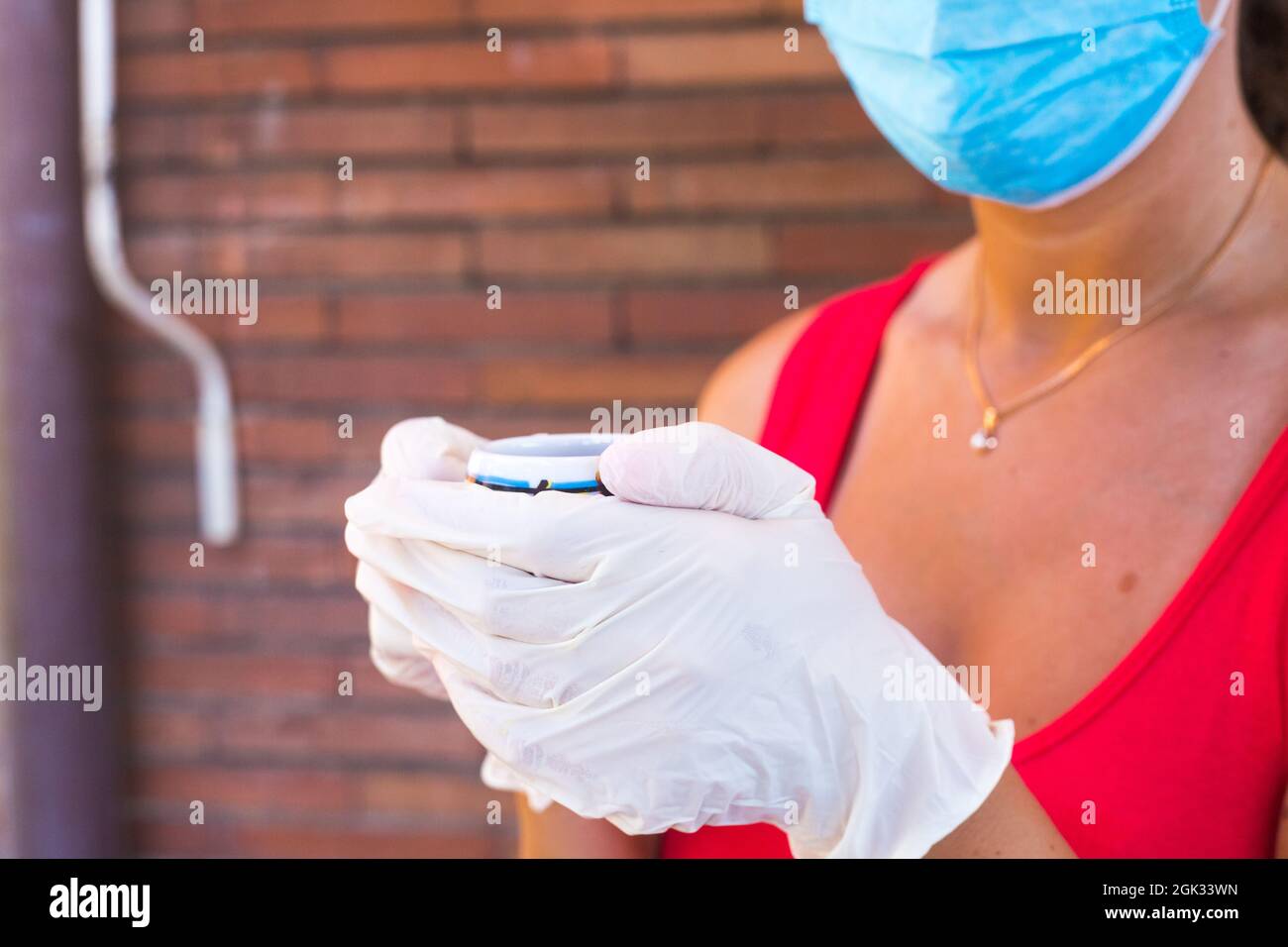 Frau in rotem Hemd, blauer handgemaskierter OP-Maske und weißen medizinischen Handschuhen trinkt Kaffee im Freien. Backsteinmauer dahinter. Kaffee nach der Pandemie mitnehmen Stockfoto