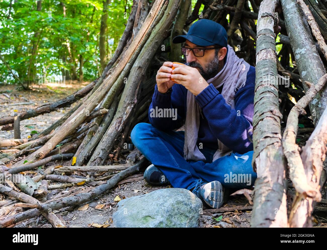 Junger Mann im Wald trinkt Kaffee unter einem Tierheim. Überleben und Camping Konzept des Lebens in der Wildnis Stockfoto
