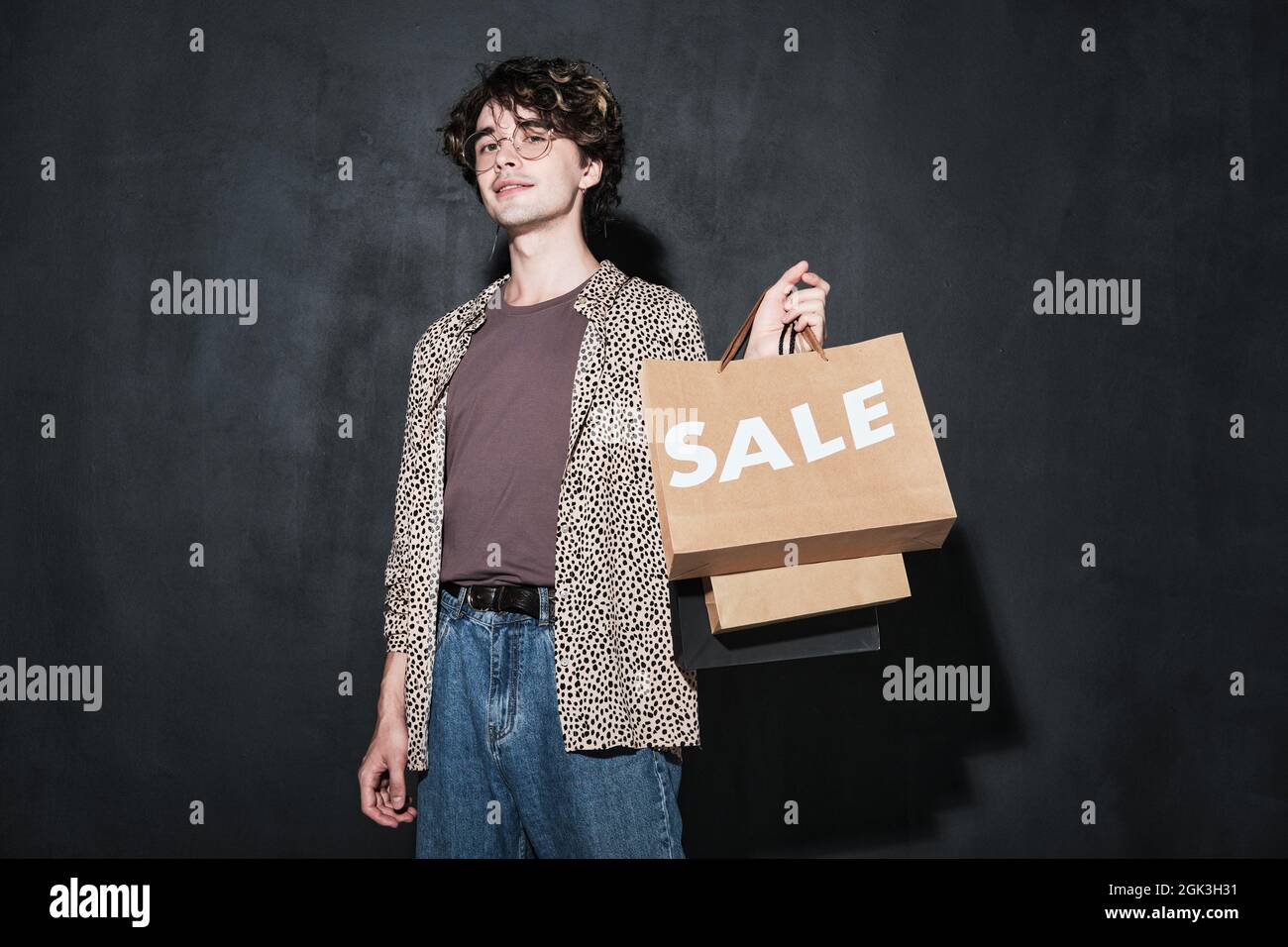 Porträt eines jungen, stilvollen Mannes mit Papiertüten, der die Kamera vor dem schwarzen Hintergrund betrachtet Stockfoto