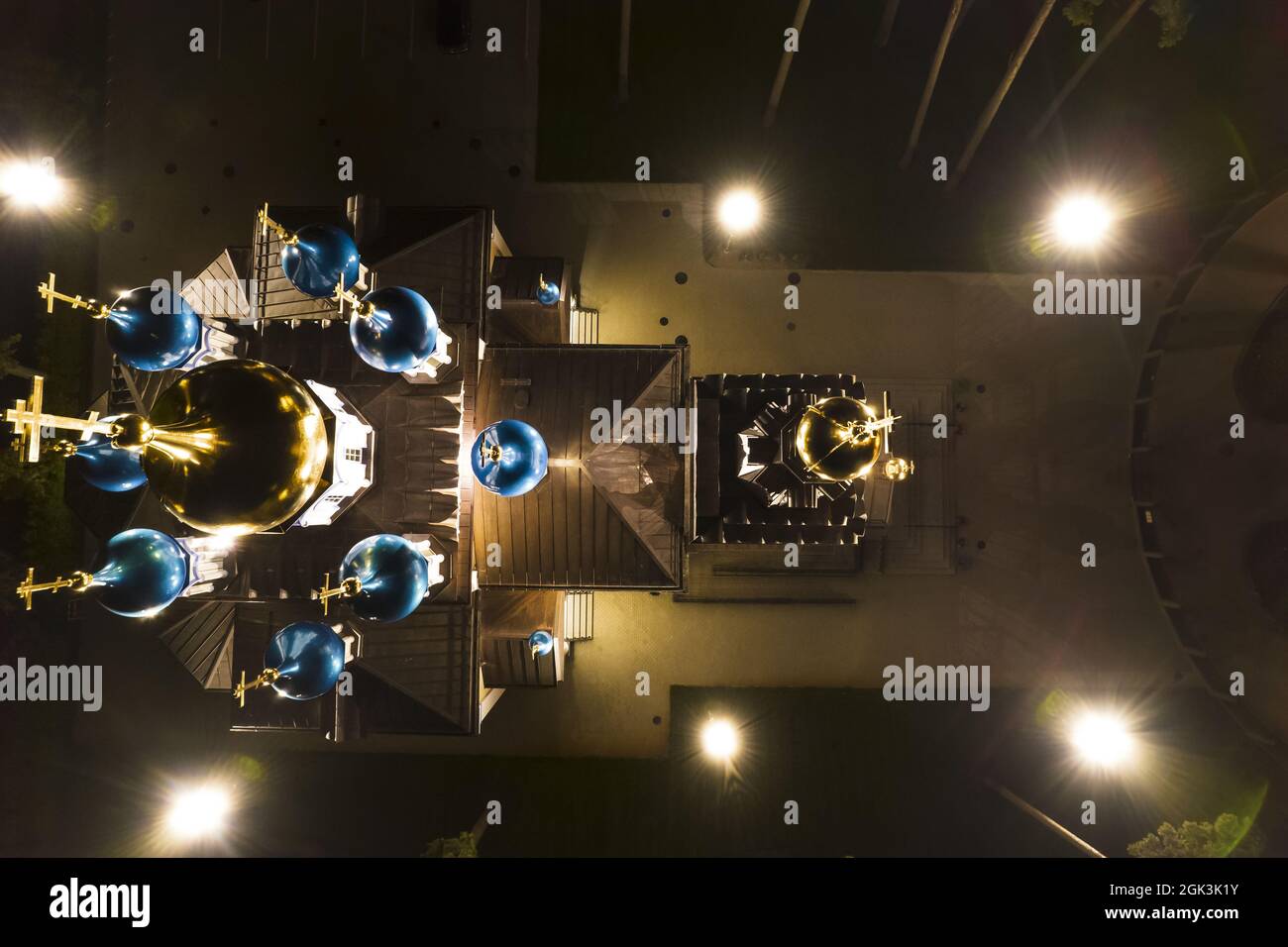 Luftaufnahme der beleuchteten Kirche zwischen Kiefern in der Nacht. Neue öffentliche leere weiße Kapelle mit goldenen und blauen Kuppeln im Freien von oben. Schöner religiöser, heiliger Ort für Ämter Stockfoto