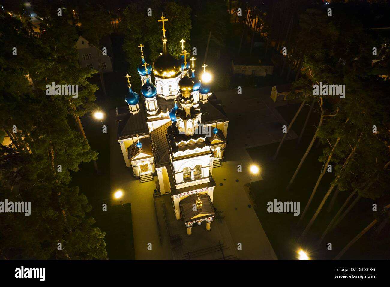 Luftaufnahme der beleuchteten Kirche zwischen Kiefern in der Nacht. Neue öffentliche leere weiße Kapelle mit goldenen und blauen Kuppeln im Freien von oben. Schöner religiöser, heiliger Ort für Ämter Stockfoto