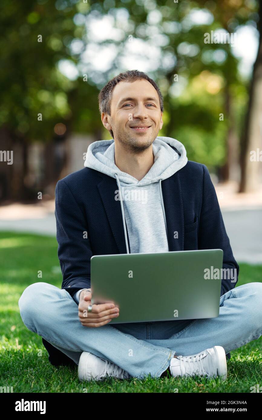 Porträt eines lächelnden Projektmanagers Fernarbeit am Computer im Park, auf dem Rasen sitzend. Distanzauftrag Stockfoto