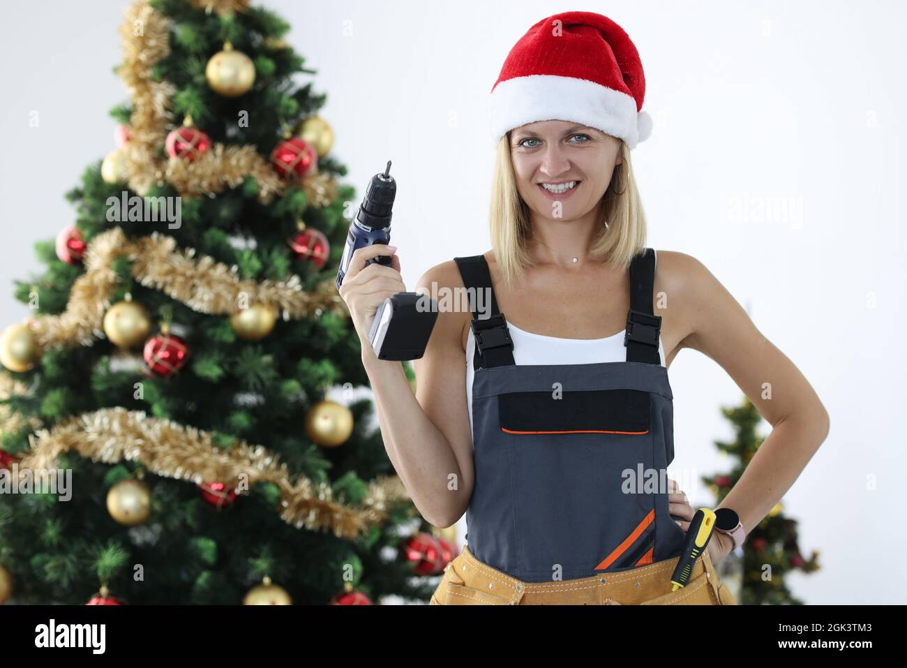 Lächelnde Frau in Uniform und weihnachtsmannhut mit Bohrmaschine in den Händen in der Nähe des weihnachtsbaums Stockfoto