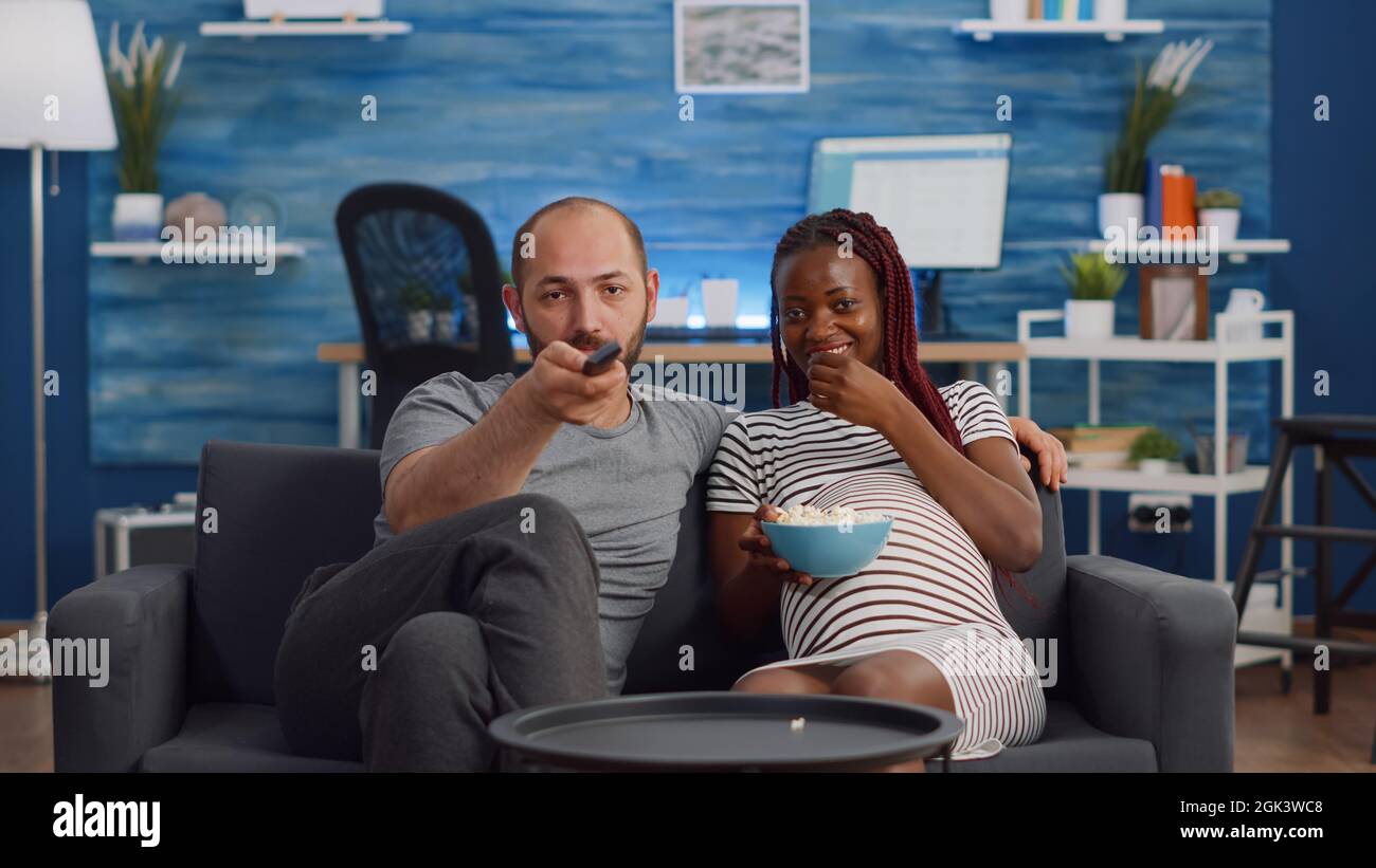 POV von interracial paar mit Schwangerschaft Fernsehen und Popcorn essen. Multiethnische Menschen, die ein Kind erwarten, sitzen auf dem Sofa. Partner, die Spaß haben, während sie die Kamera betrachten Stockfoto