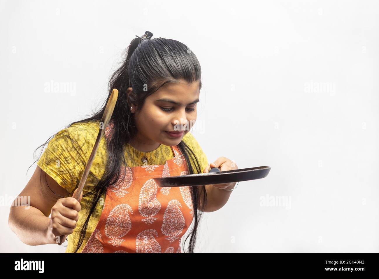 Eine hübsche indische Hausfrau in Kochschürze mit Holzspatel und Bratpfanne in den Händen auf weißem Hintergrund Stockfoto