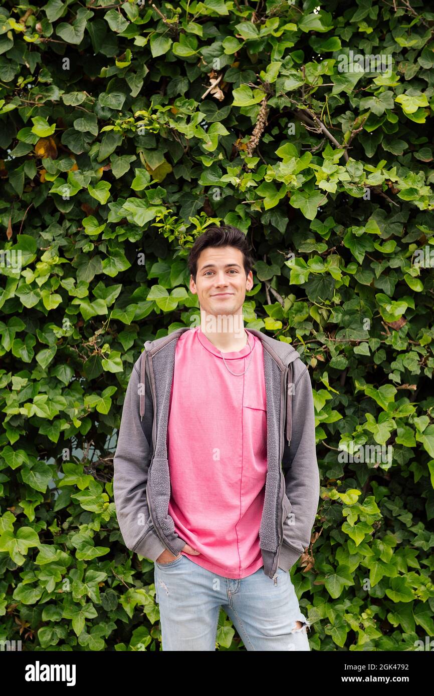 Vertikales Foto eines freundlichen Teenagers, der lächelnd die Kamera auf einem grünen Pflanzenhintergrund anschaut, Konzept menschlicher Ausdrücke und Emotionen, Kopierraum fo Stockfoto